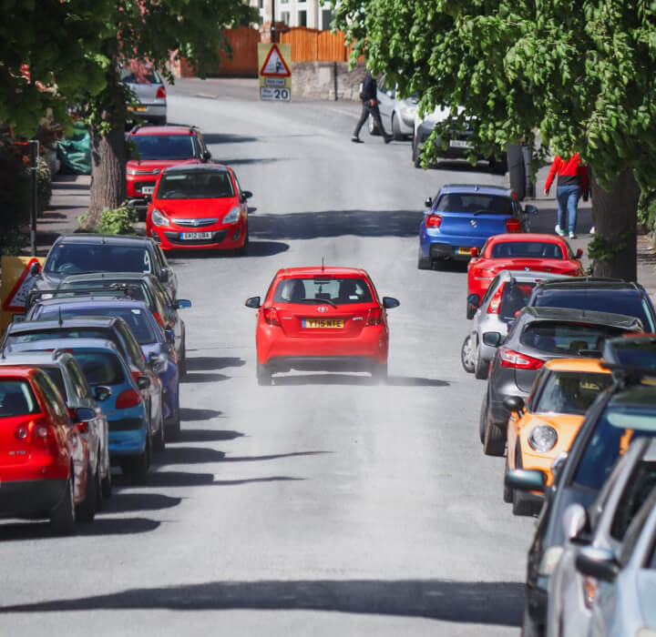 A red car driving down a residential street lined with parked cars